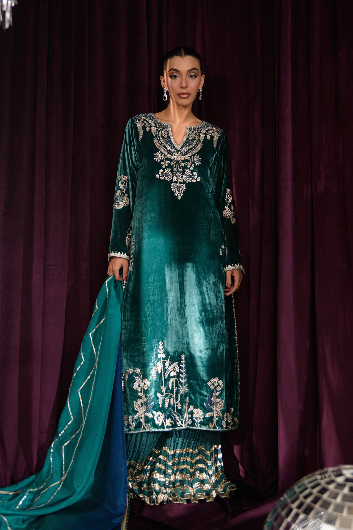 Model wearing an embroidered emerald green velvet kurta set with a matching gharara and teal dupatta.
