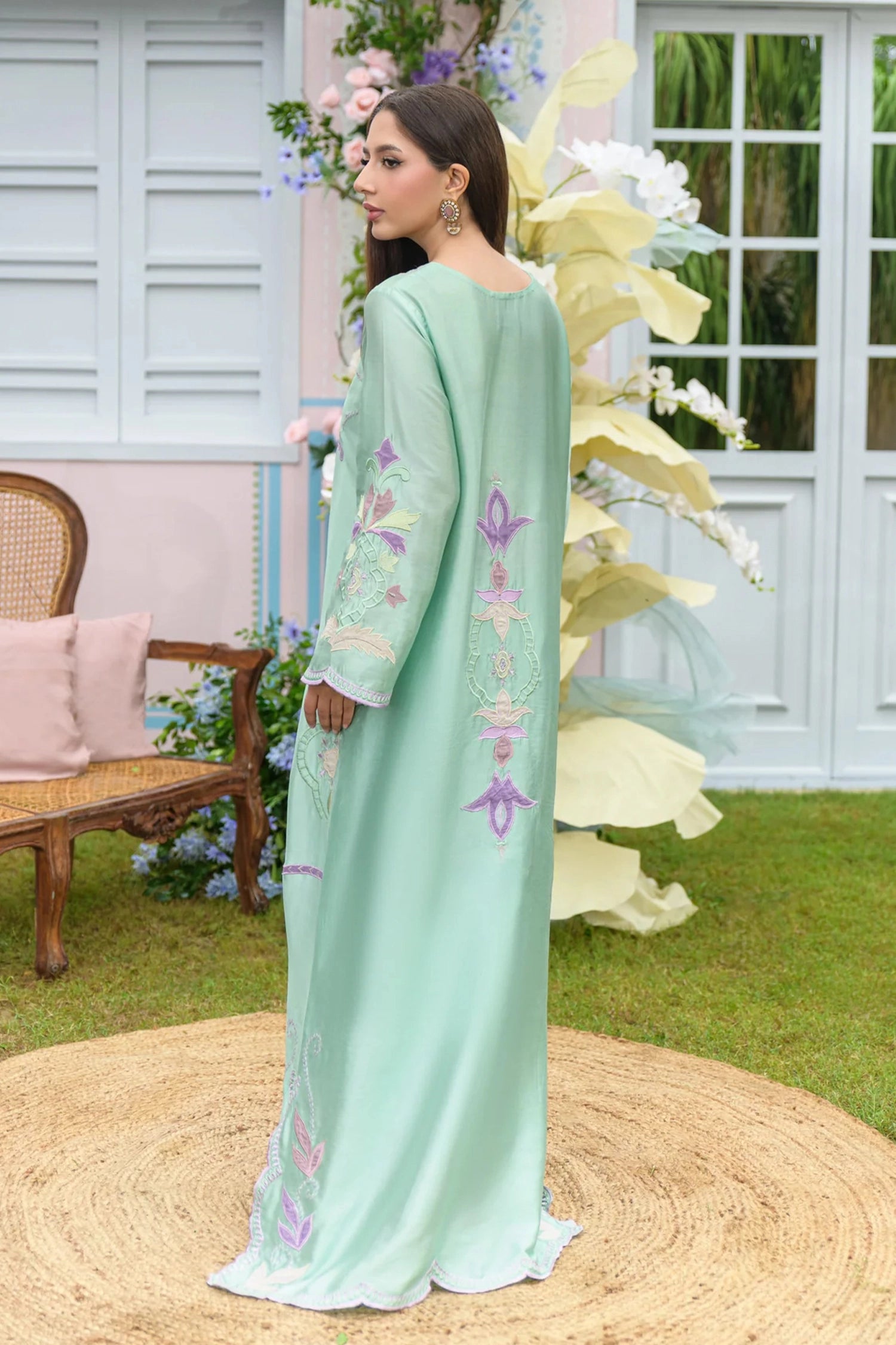 A woman modeling a long mint green traditional dress with floral embroidery on the sleeves and side, standing in an outdoor garden setting with a woven rug underfoot.

