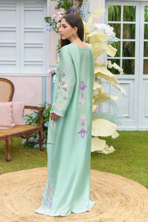 A woman modeling a long mint green traditional dress with floral embroidery on the sleeves and side, standing in an outdoor garden setting with a woven rug underfoot.

