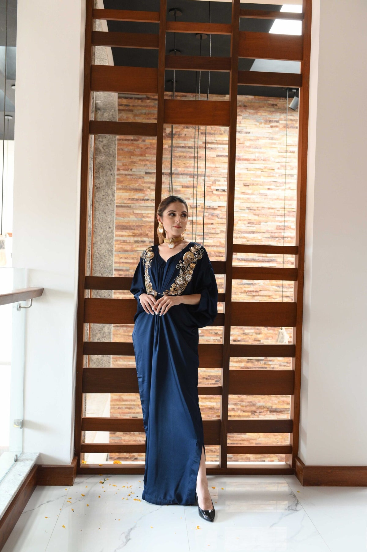Model wearing a navy blue satin kaftan dress with gold floral chest embroidery and a side slit.
