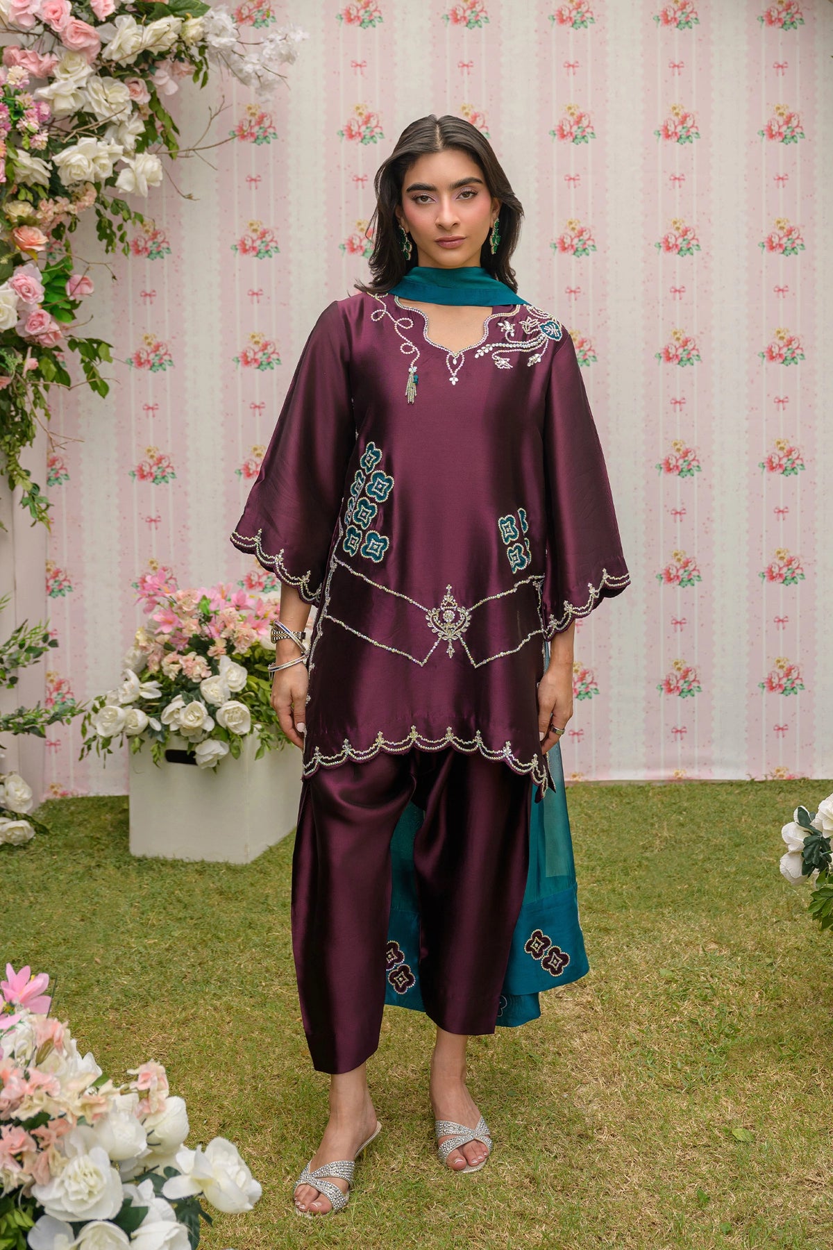Woman in a burgundy and teal outfit standing in front of floral decorations.

