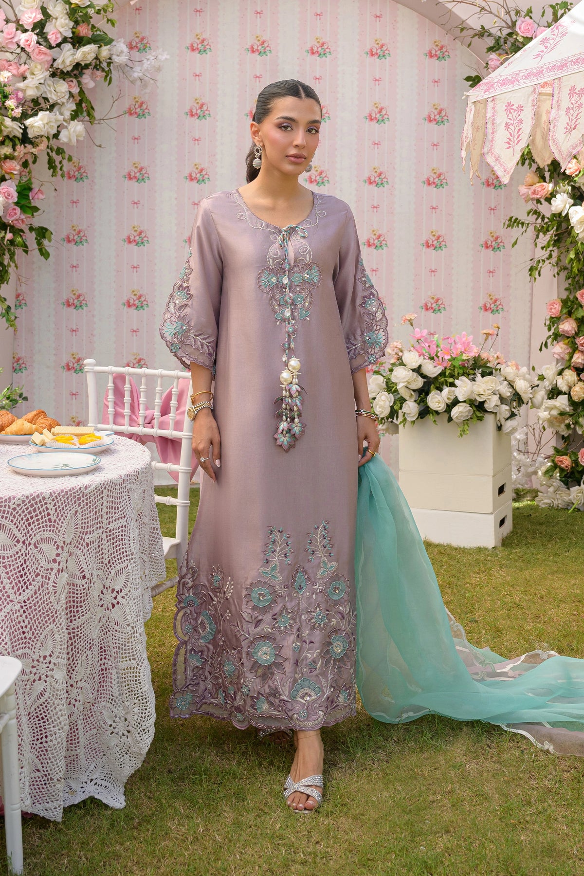 Woman in a lavender dress with floral embroidery standing outdoors with floral decorations and tables.

