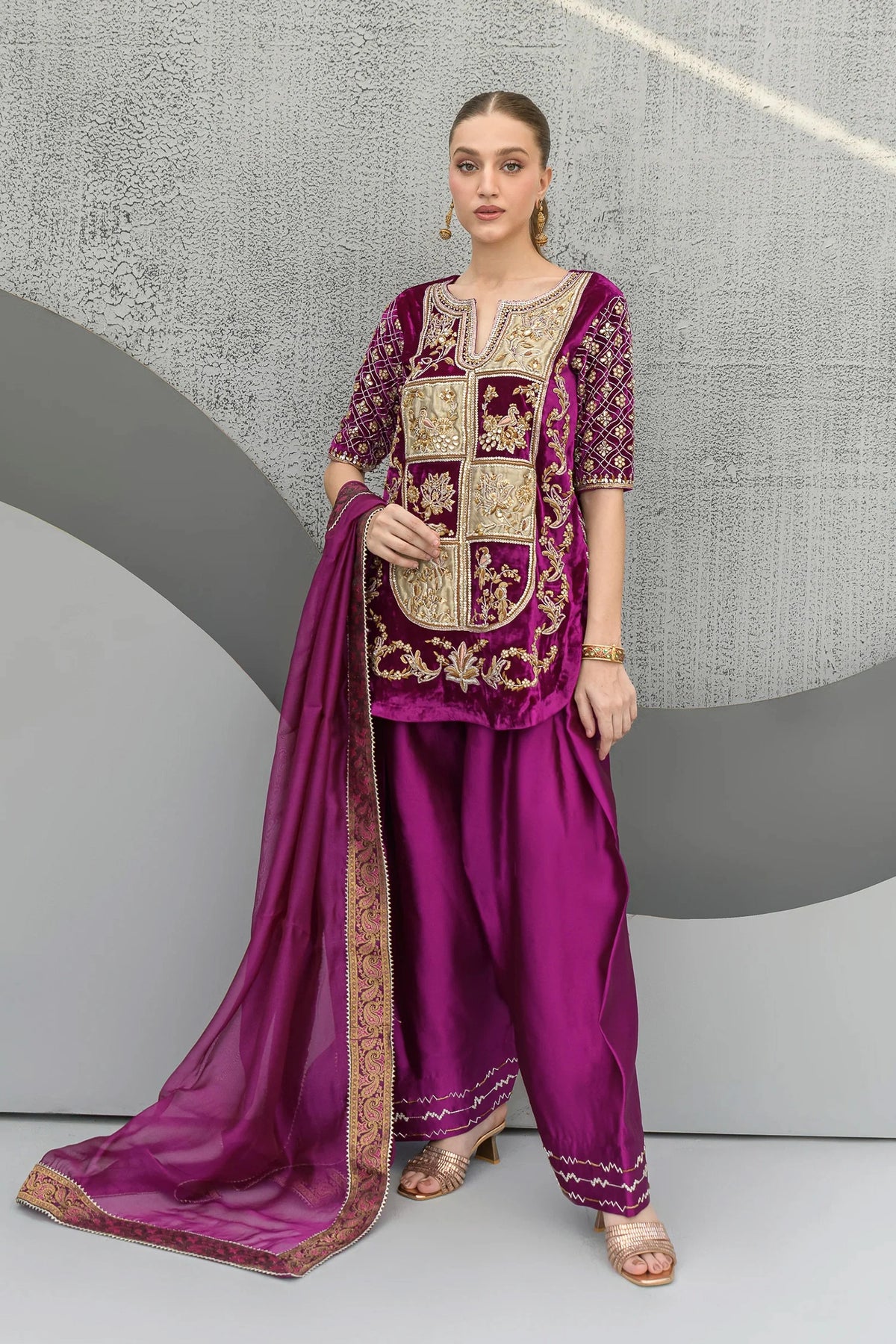 Woman wearing a traditional purple outfit with gold embroidery against a gray background

